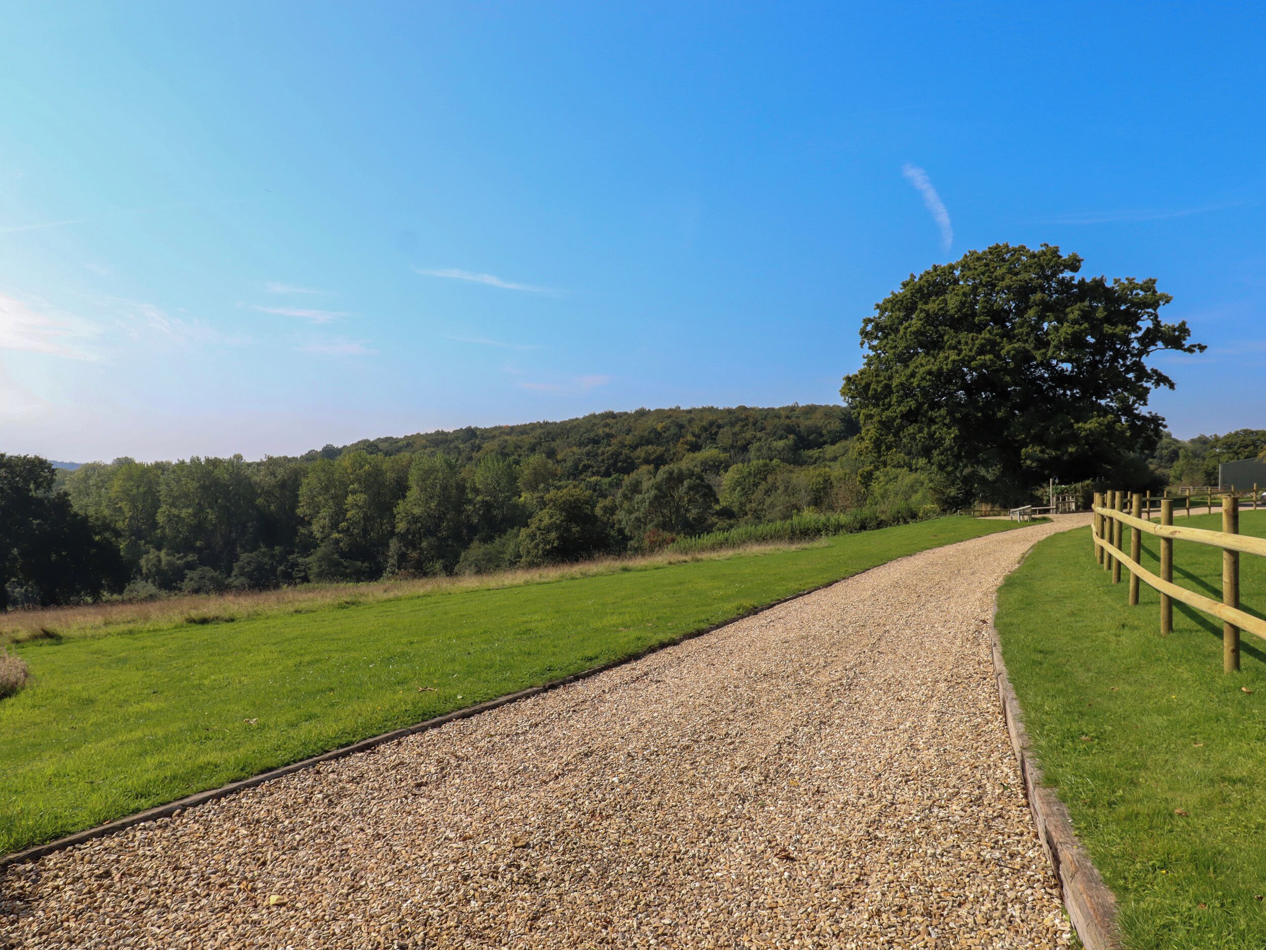 A gravel path leading through a green field with a wooden fence on one side and a large tree on the other.