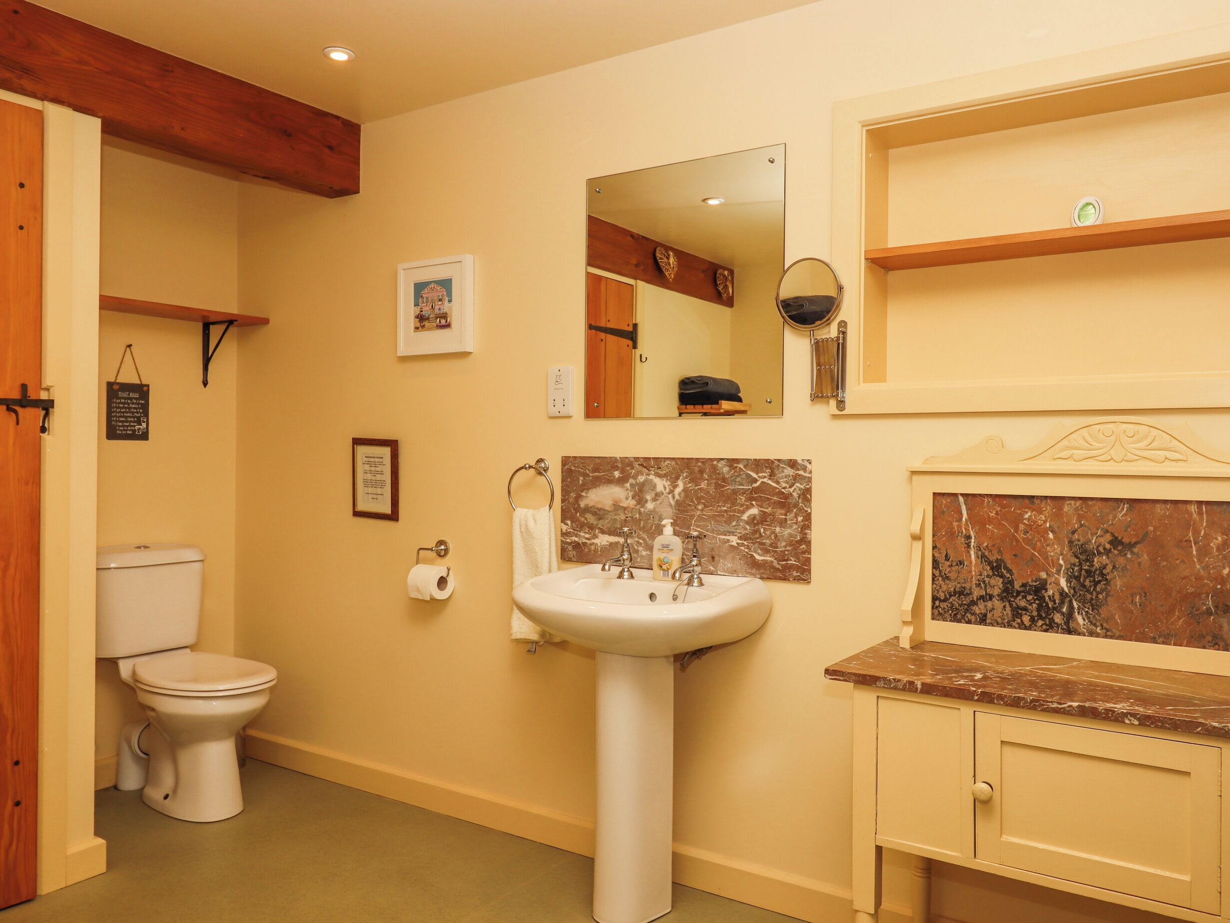 Ein Badezimmer mit Toilette, Waschbecken und Spiegel. Über dem Waschbecken befindet sich ein Regal und an der Wand ein gerahmtes Bild.