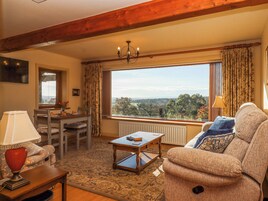 A cozy living room with a wooden ceiling, a dining table with chairs, a sofa, a lamp, and a view of the outdoors.