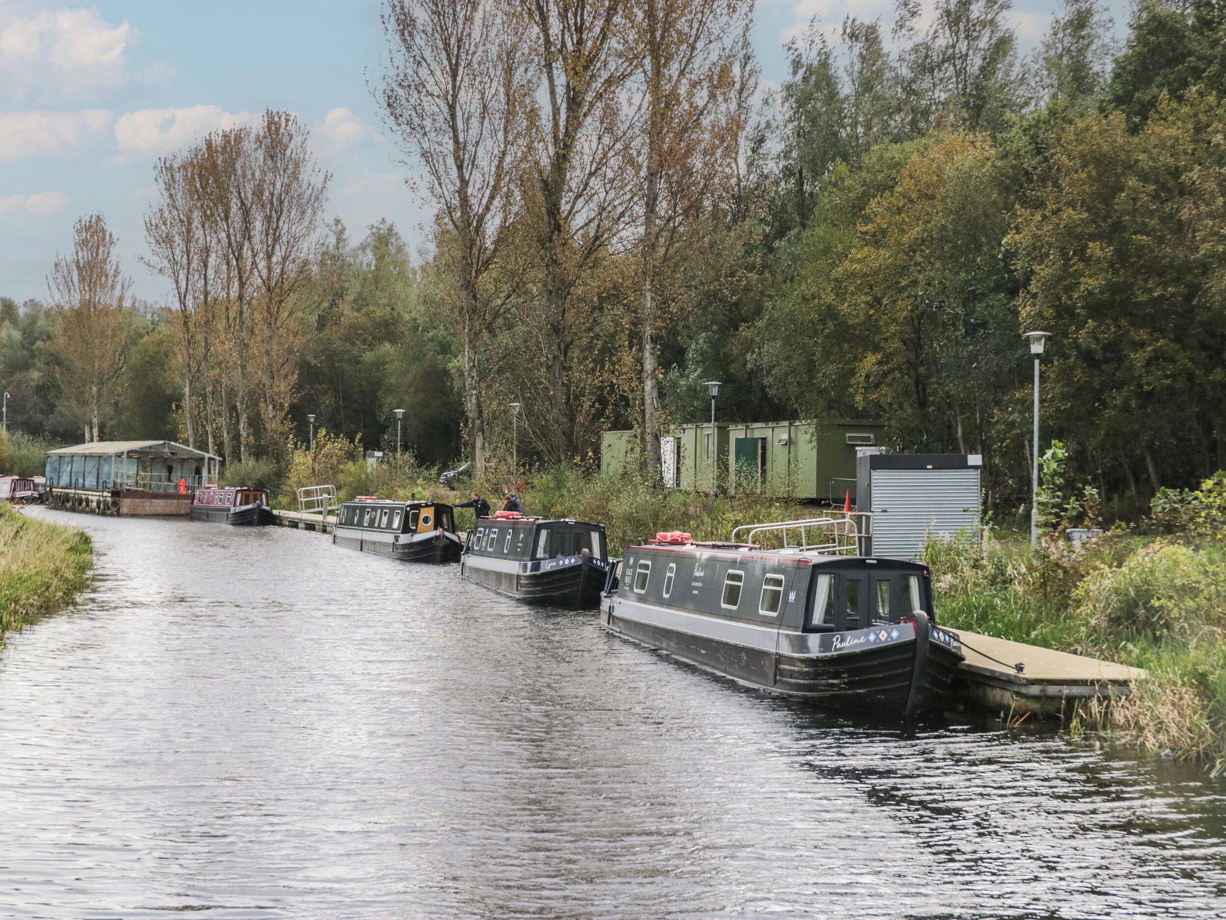 A narrow canal with several narrowboats docked along the sides. There are trees and greenery on both sides of the canal, and a small building is visible on the right side.