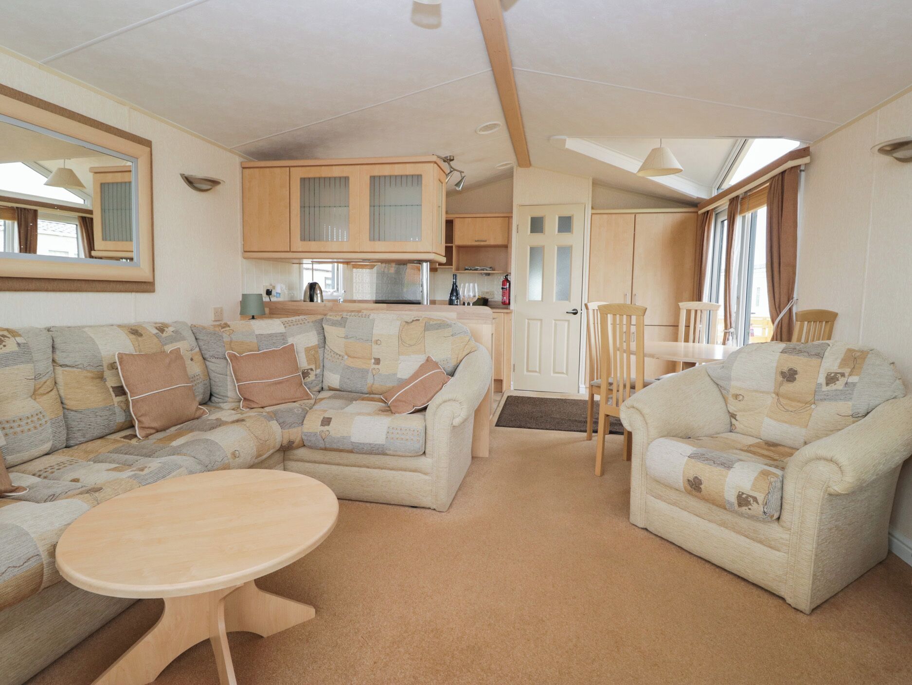 A cozy living room with a sectional sofa, a round coffee table, and a dining area with a small table and chairs. The room features wooden cabinets and a large mirror on the wall.