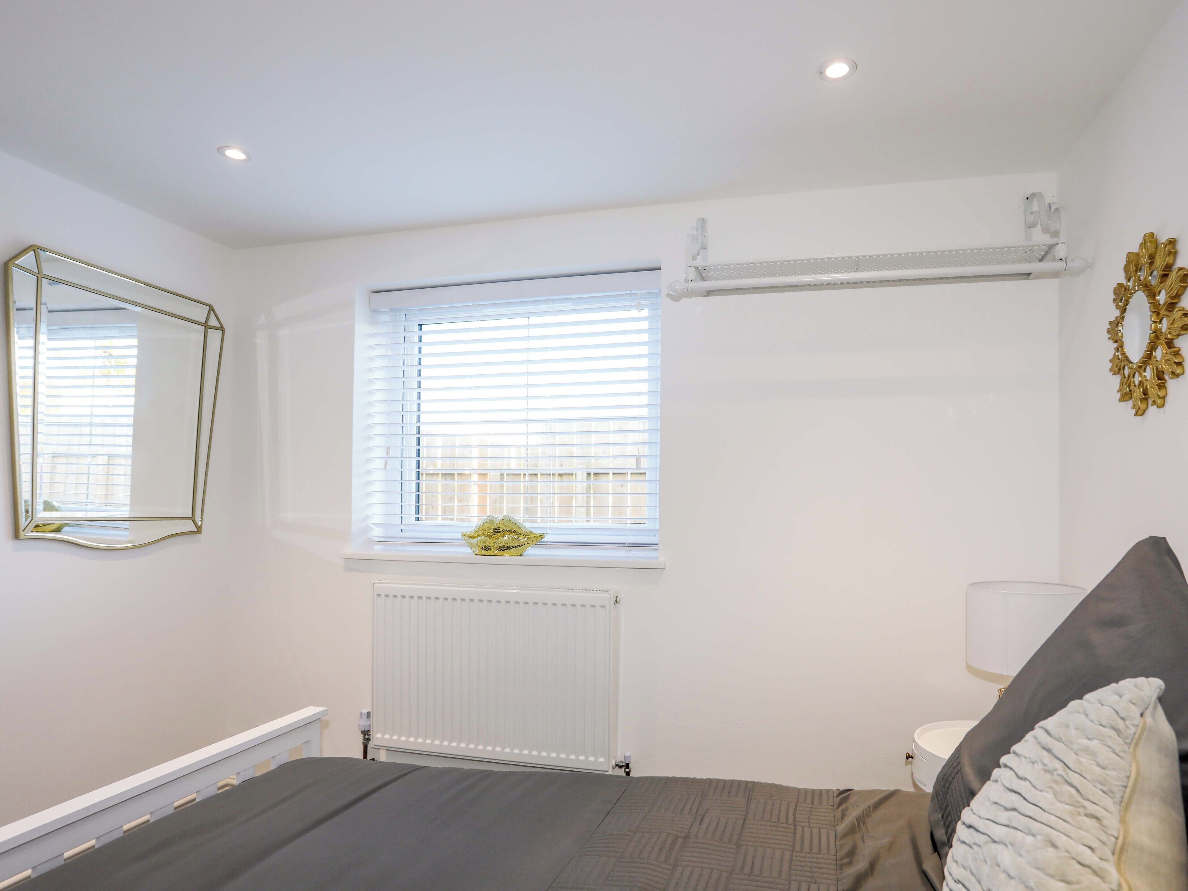A bedroom with a bed, a window with blinds, a radiator, a mirror, and a wall-mounted light fixture.