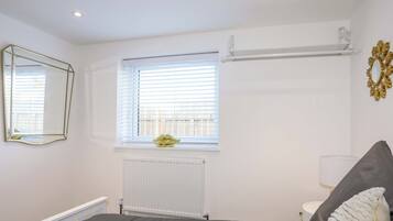 A bedroom with a bed, a window with blinds, a radiator, a mirror, and a wall-mounted light fixture.