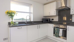 A modern kitchen with white cabinets, a stainless steel range hood, a microwave, and a window with blinds. - Ty'n Ffridd Bach (Pwllheli)