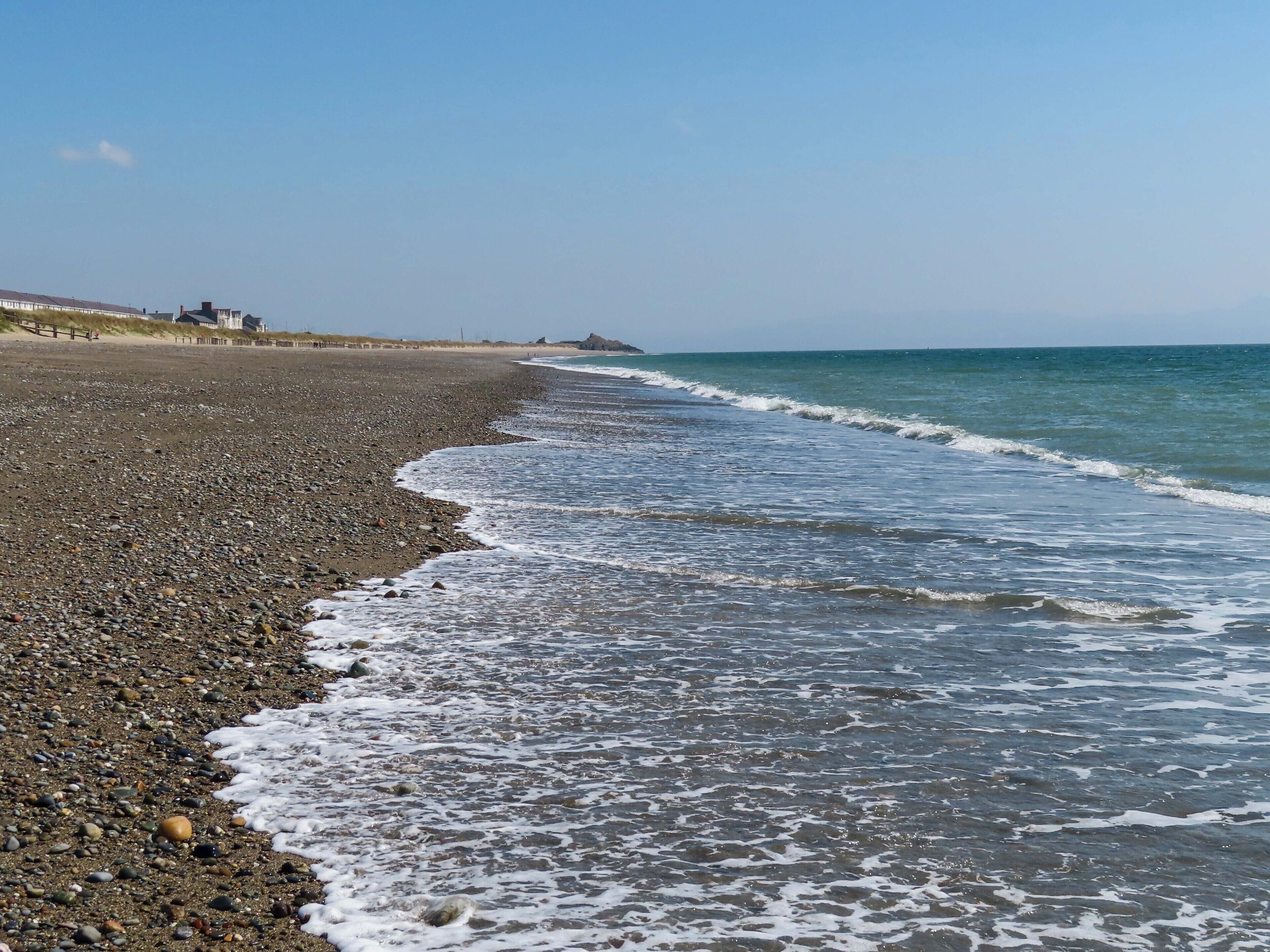 A pebbled beach with waves lapping at the shore, a clear sky, and buildings in the distance.