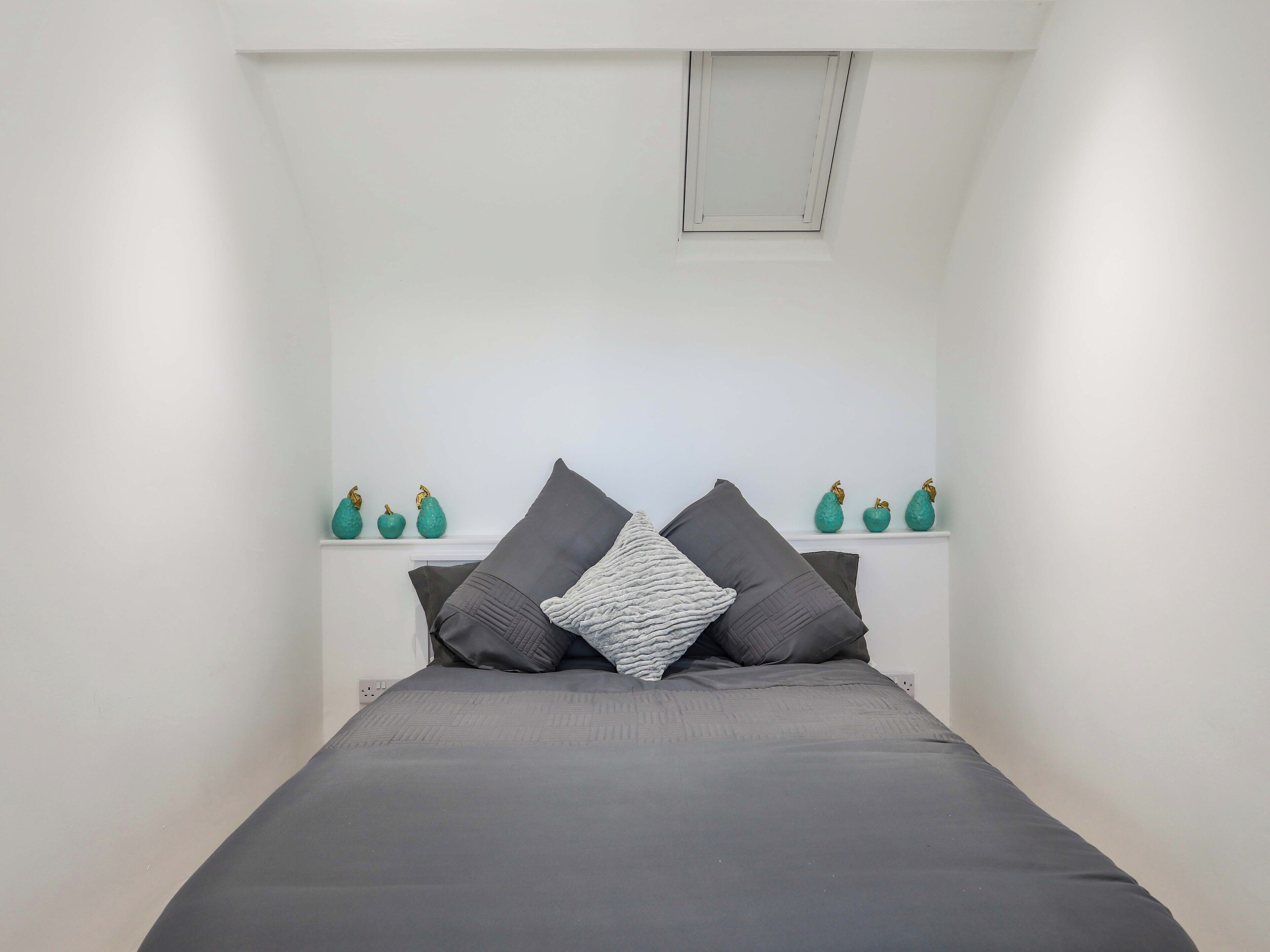 Ein kleiner, weißer Raum mit einem Bett, zwei Kissen und einem Regal mit Dekoration.