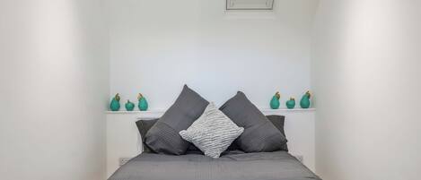 Ein kleiner, weißer Raum mit einem Bett, zwei Kissen und einem Regal mit Dekoration.