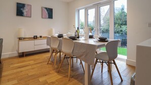 A modern dining area with a wooden table, white chairs, and a large window offering a view of a garden. - 3 Grange Villas (Bridport)