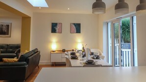 A modern dining area with a table set for two, overhead pendant lighting, and a large window with a view of greenery. - 3 Grange Villas (Bridport)