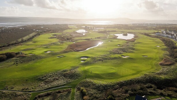 Vista aérea de un campo de golf con varios hoyos, obstáculos acuáticos y una costa distante.