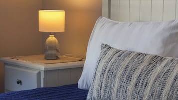 Una cama bien tendida con una manta azul y un cojín a rayas, una lámpara de noche y una mesita de noche blanca.