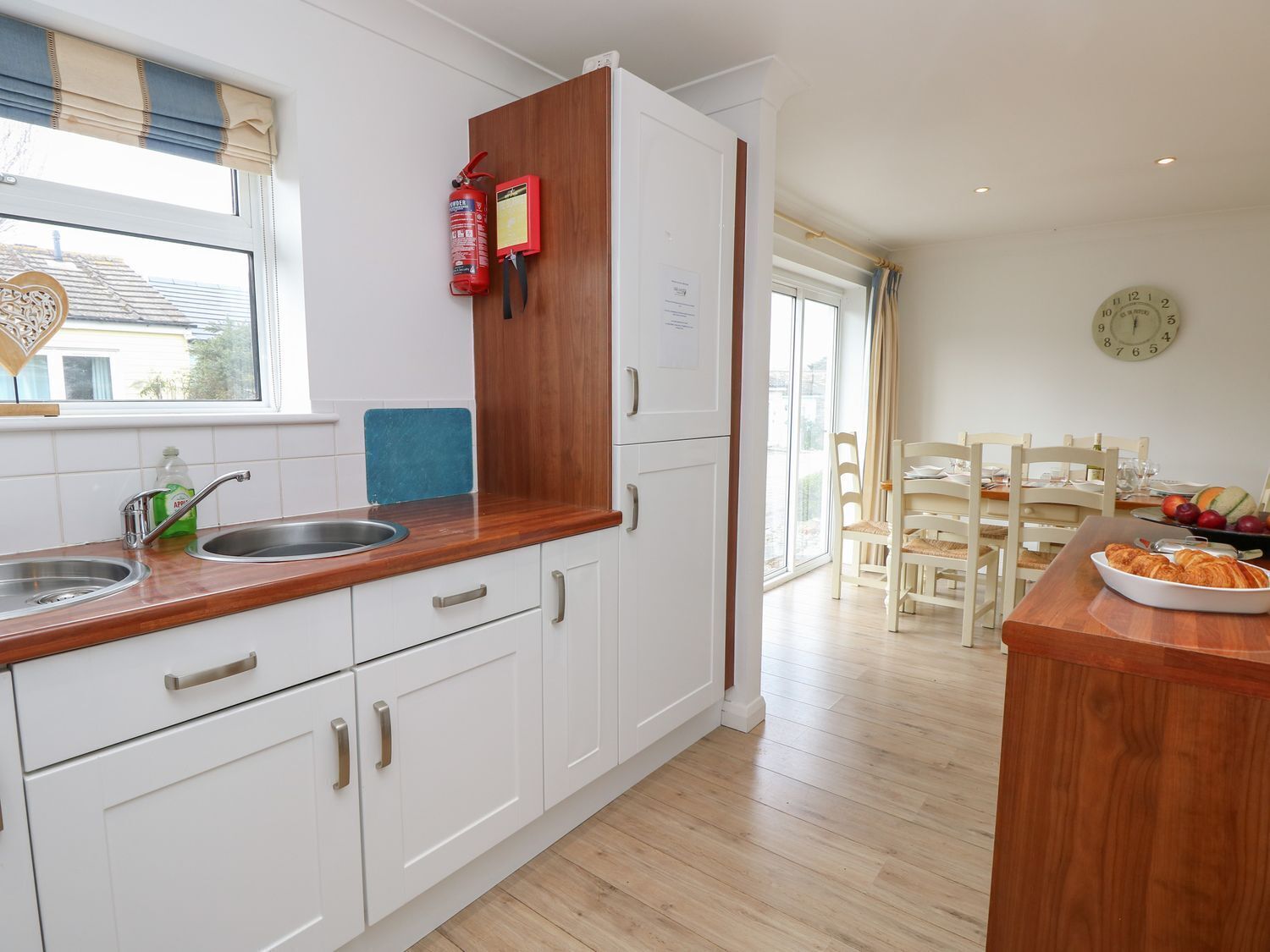 A modern kitchen with white cabinets, a wooden countertop, and a built-in oven. There is a window with a view of the outside, a clock on the wall, and a dining area with a table and chairs.