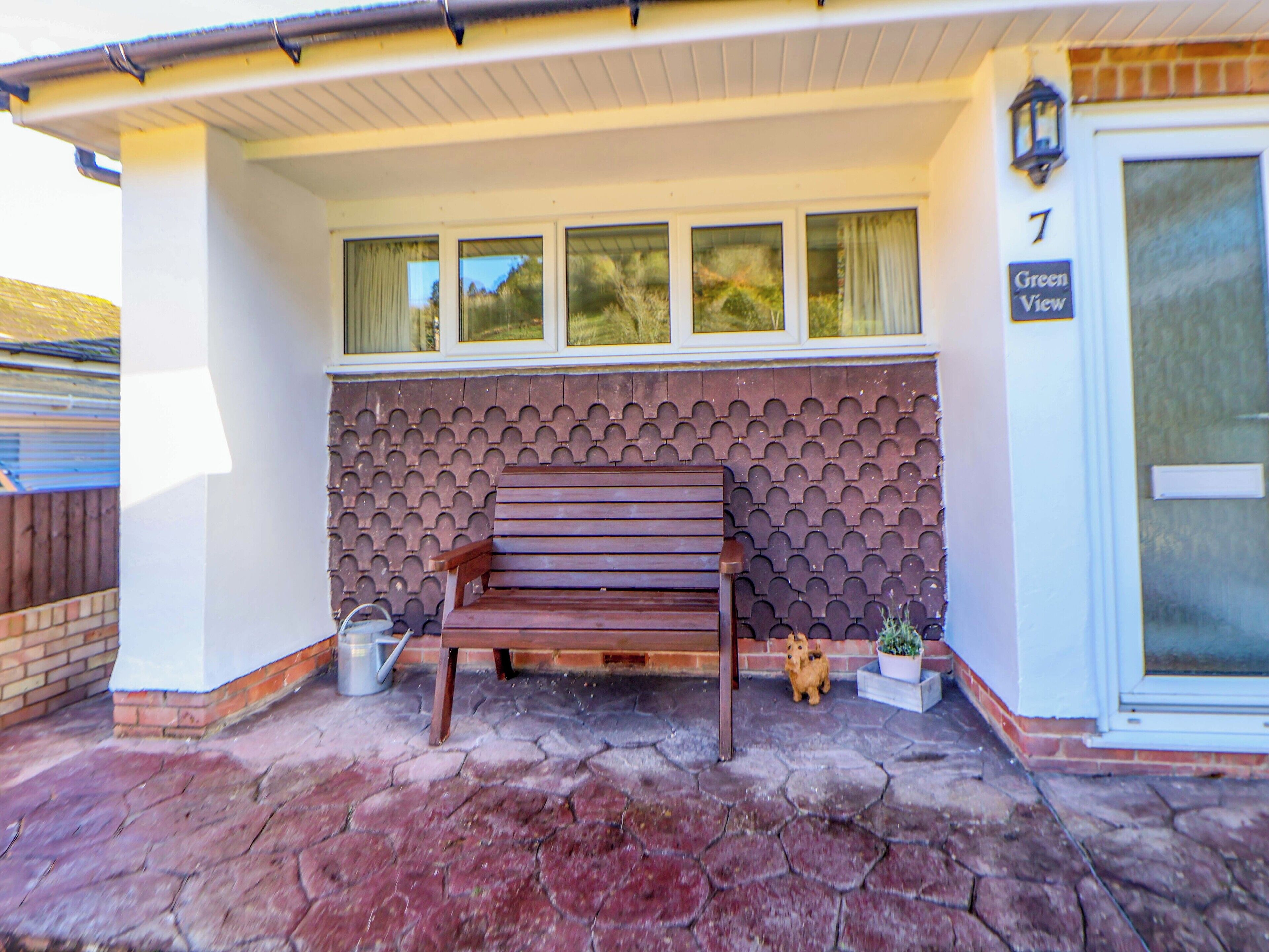 A wooden bench outside a house with a brick wall and a window.