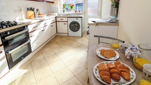 A modern kitchen with white cabinets, a gas stove, an oven, a washing machine, and a dining area with breakfast items. - Green View (Llangollen)