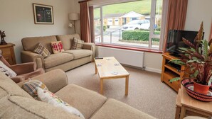 A living room with a sofa, armchair, coffee table, and a TV on a wooden stand. - Green View (Llangollen)