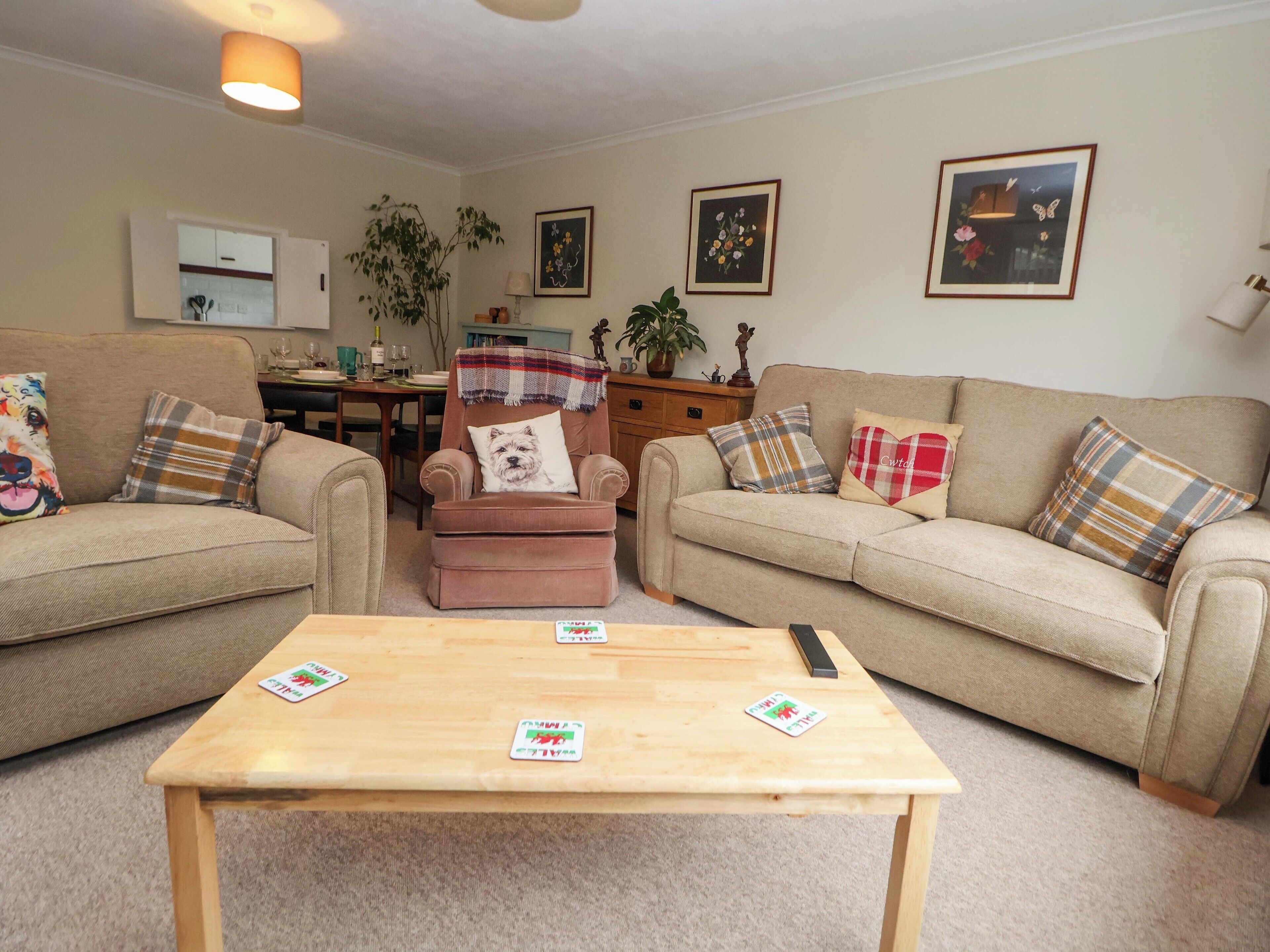A living room with two sofas, a wooden coffee table, and a small armchair with a dog pillow. There are framed pictures on the walls and a plant in the corner.