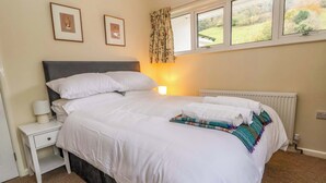 A neatly made bed with white linens, a bedside table with a lamp, and a window with curtains. - Green View (Llangollen)