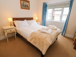 A neatly made bed with white linens and a beige throw blanket, two bedside lamps, a nightstand with a drawer, and a window with blue curtains. - Garden Retreat (Swanage)