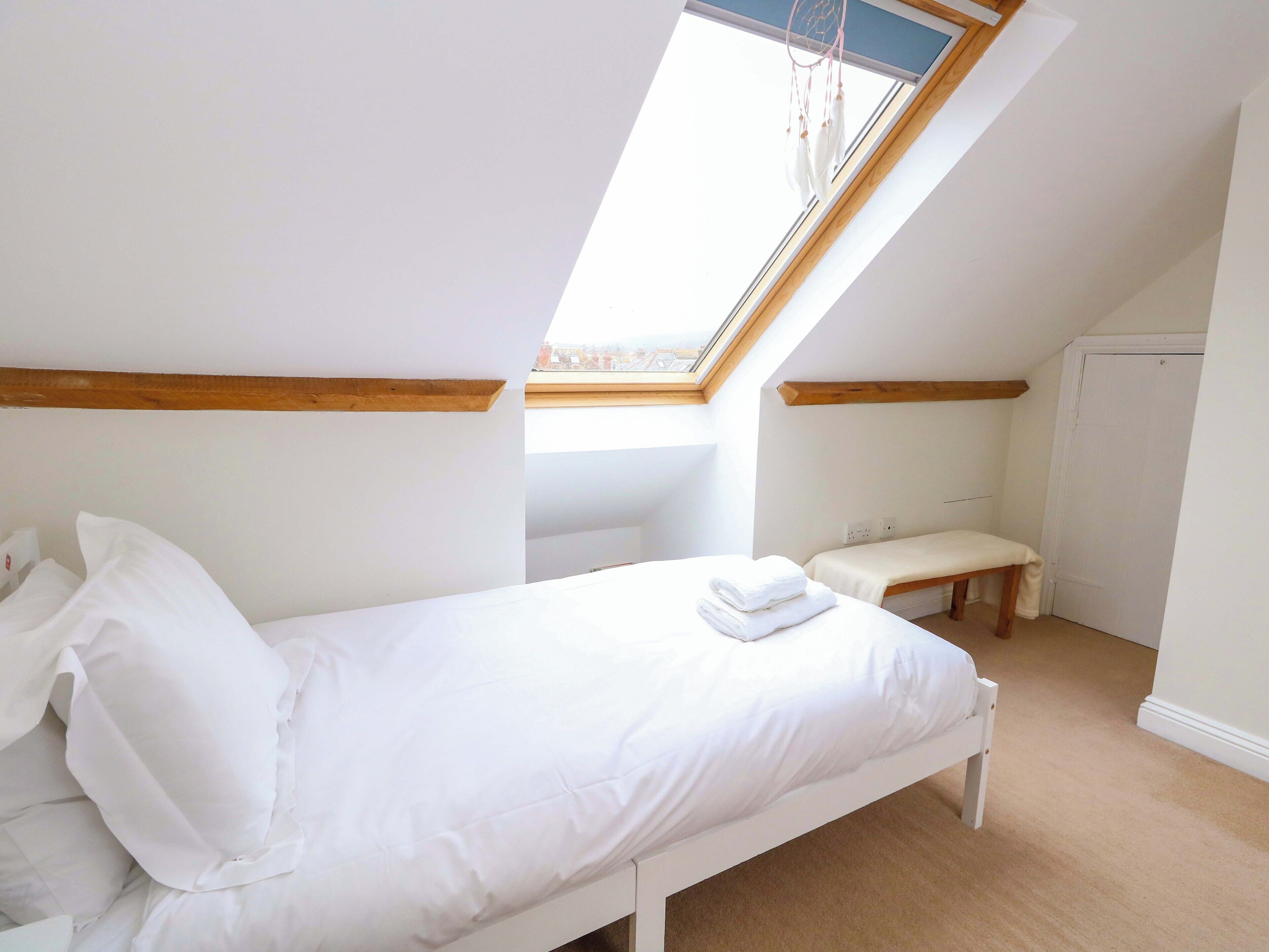 A small, white, single-bed room with a skylight, wooden beams, and a bench.