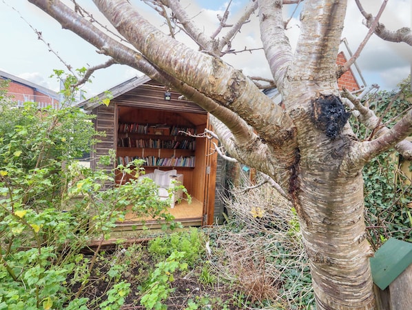 A wooden structure with a bookshelf filled with books, surrounded by greenery and a tree.