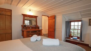 A bedroom with a bed, a wooden dresser with a mirror, a window with curtains, and a door. - Rose Cottage (Falmouth)