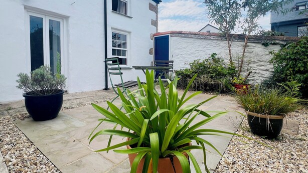 A white house with a tiled patio, a table, and chairs, surrounded by potted plants and a view of a river. - Rose Cottage (Falmouth)