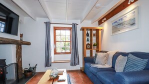 A cozy living room with a blue sofa, a wooden coffee table, a fireplace, and a window with curtains. - Rose Cottage (Falmouth)