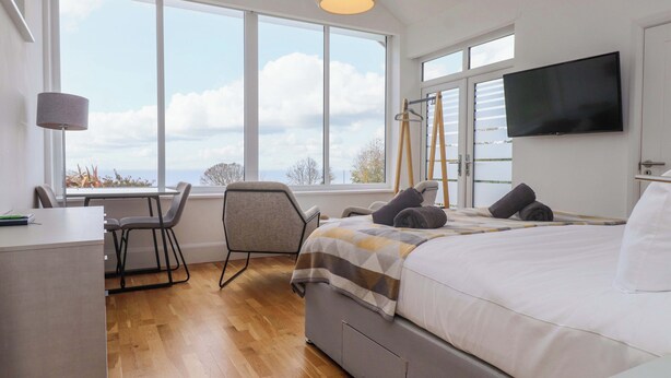 A modern bedroom with a large bed, a desk, and a chair. There are large windows offering a view of the sky and clouds.