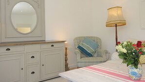 A room with a large mirror, a dresser with drawers, a chair with a cushion, a table with a vase of flowers, and a floor lamp. - Trestar (Wadebridge)