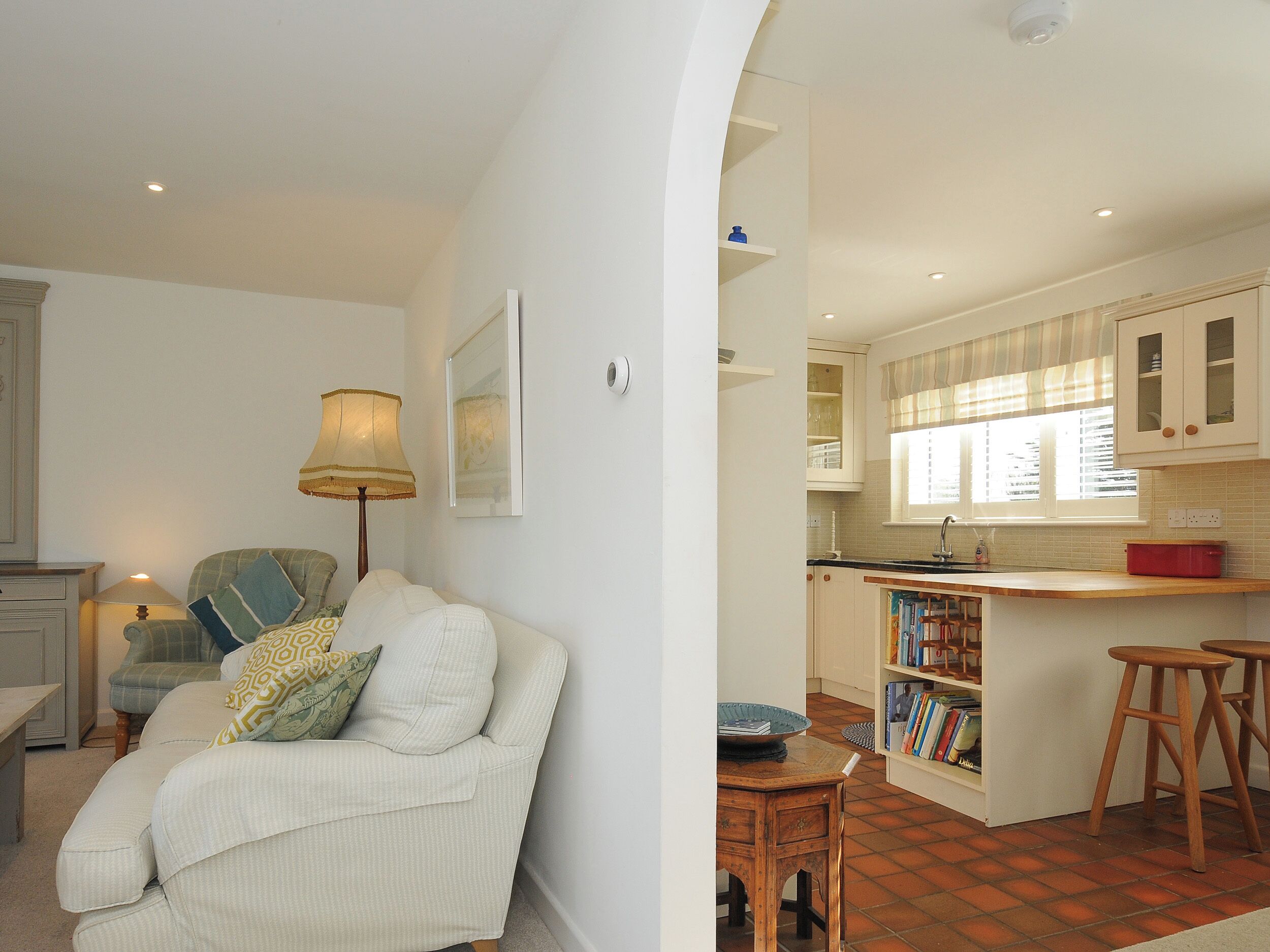 A living room with a sofa, a lamp, and a bookshelf. The kitchen has wooden cabinets, a window, and a red pot on the counter.