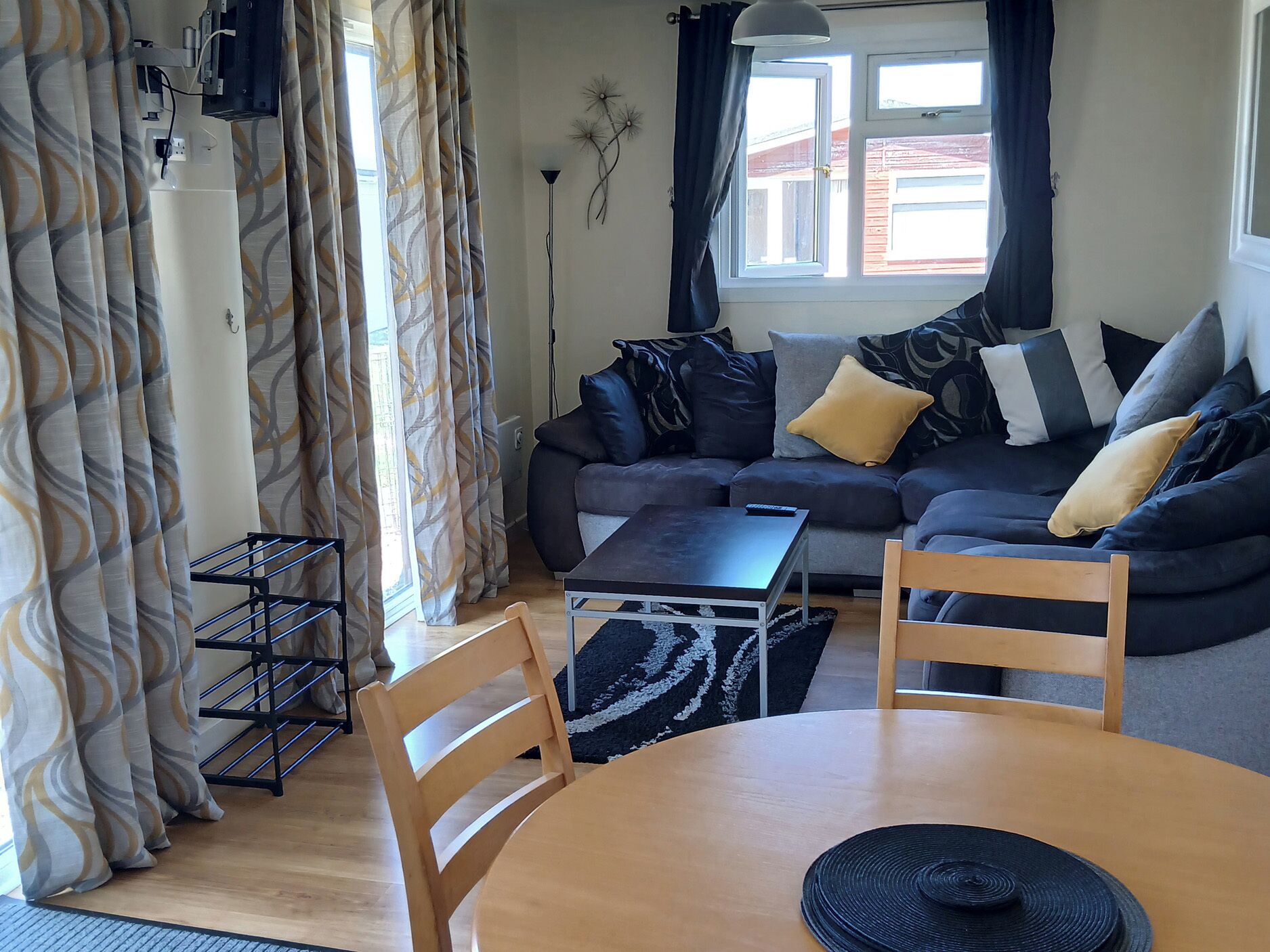 Un salon comprenant un canapé, une table basse et deux fauteuils. On y trouve une fenêtre avec des rideaux, une lampe et un élément décoratif fixé au mur.