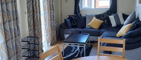 A living room with a sofa, a coffee table, and two chairs. There is a window with curtains, a lamp, and a wall-mounted decoration.