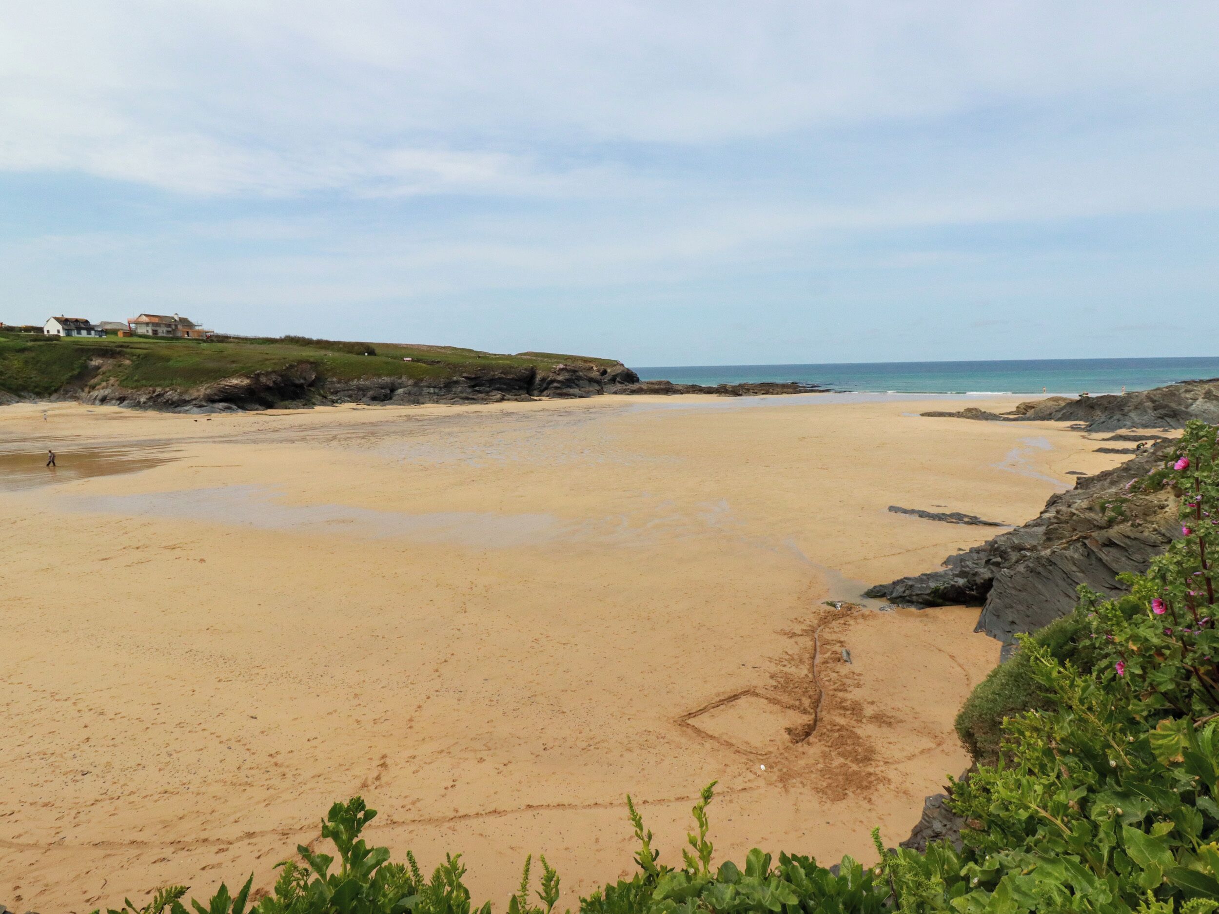 A wide sandy beach with rocky outcrops on the right, a clear sky, and houses in the background.