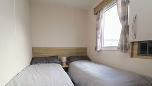 A small, simple room with two single beds, a wooden headboard, a bedside table with a lamp, and a window with curtains. - Herrylock (Rhyl)