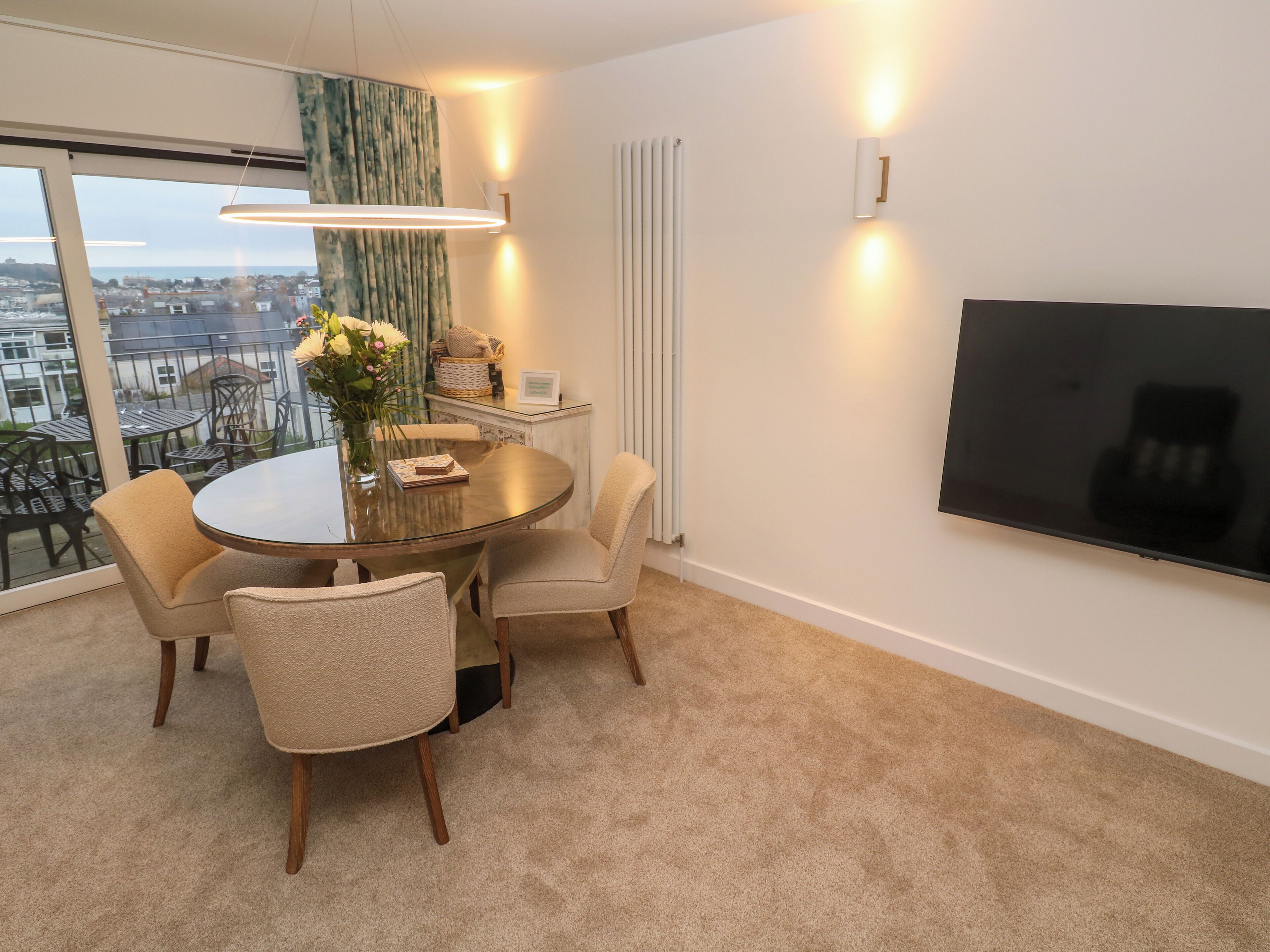 Un espace repas moderne avec une table ronde, quatre chaises, un vase rempli de fleurs, un téléviseur à écran plat fixé au mur et une vue sur la ville grâce à de grandes fenêtres.