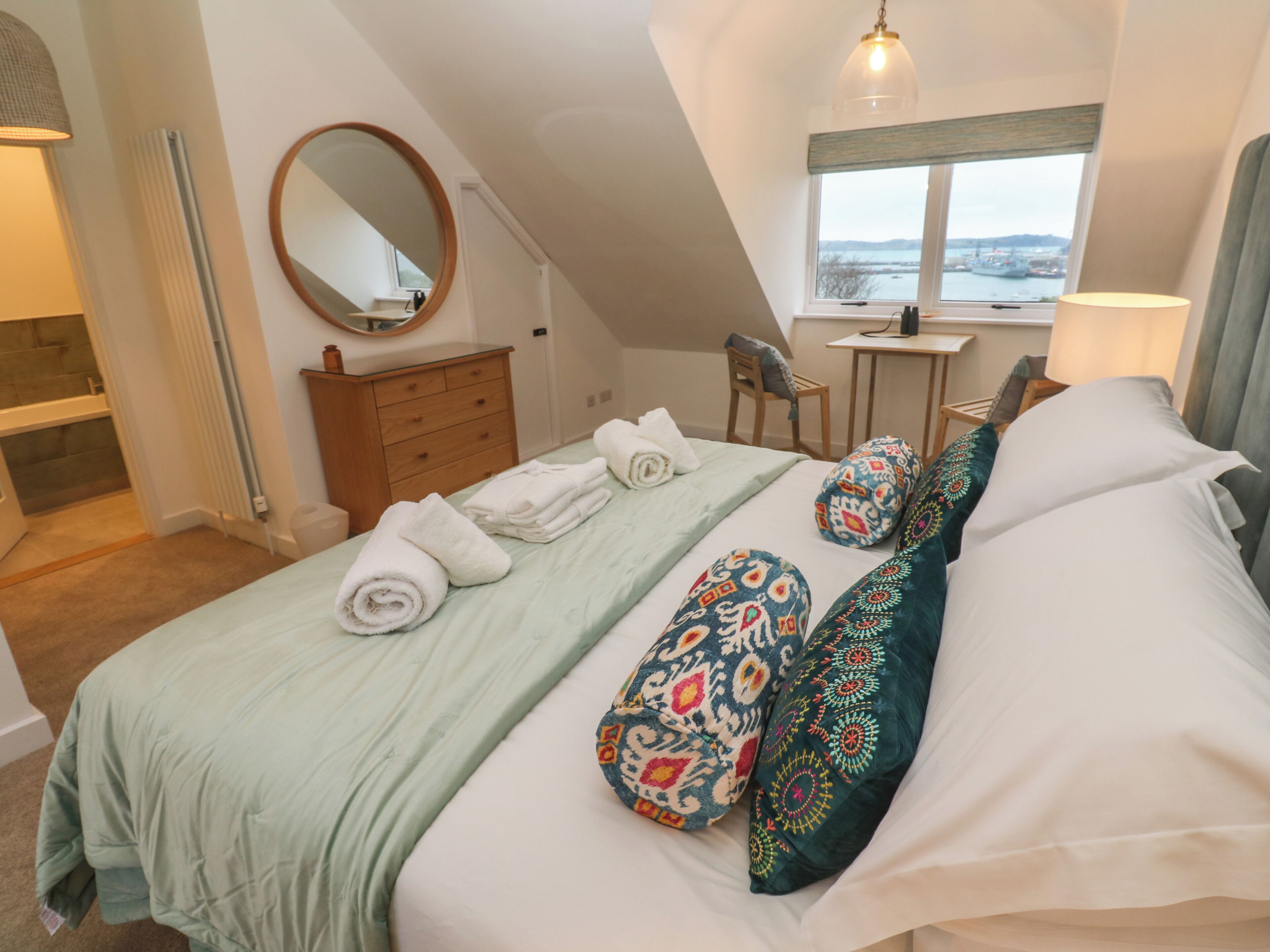 A bedroom with a bed, a dresser, a mirror, a window with a view, and a desk with a chair.