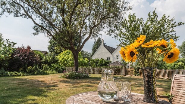 Une table ronde en bois, ornée d’un vase rempli de tournesols et d’une carafe d’eau, dans un décor de jardin.