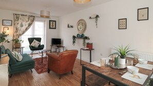 Ein modernes Wohnzimmer mit Holzboden, einem grünen Sessel, einem braunen Ledersessel, einem Esstisch mit weißen Platzsets und einer Hängeleuchte.