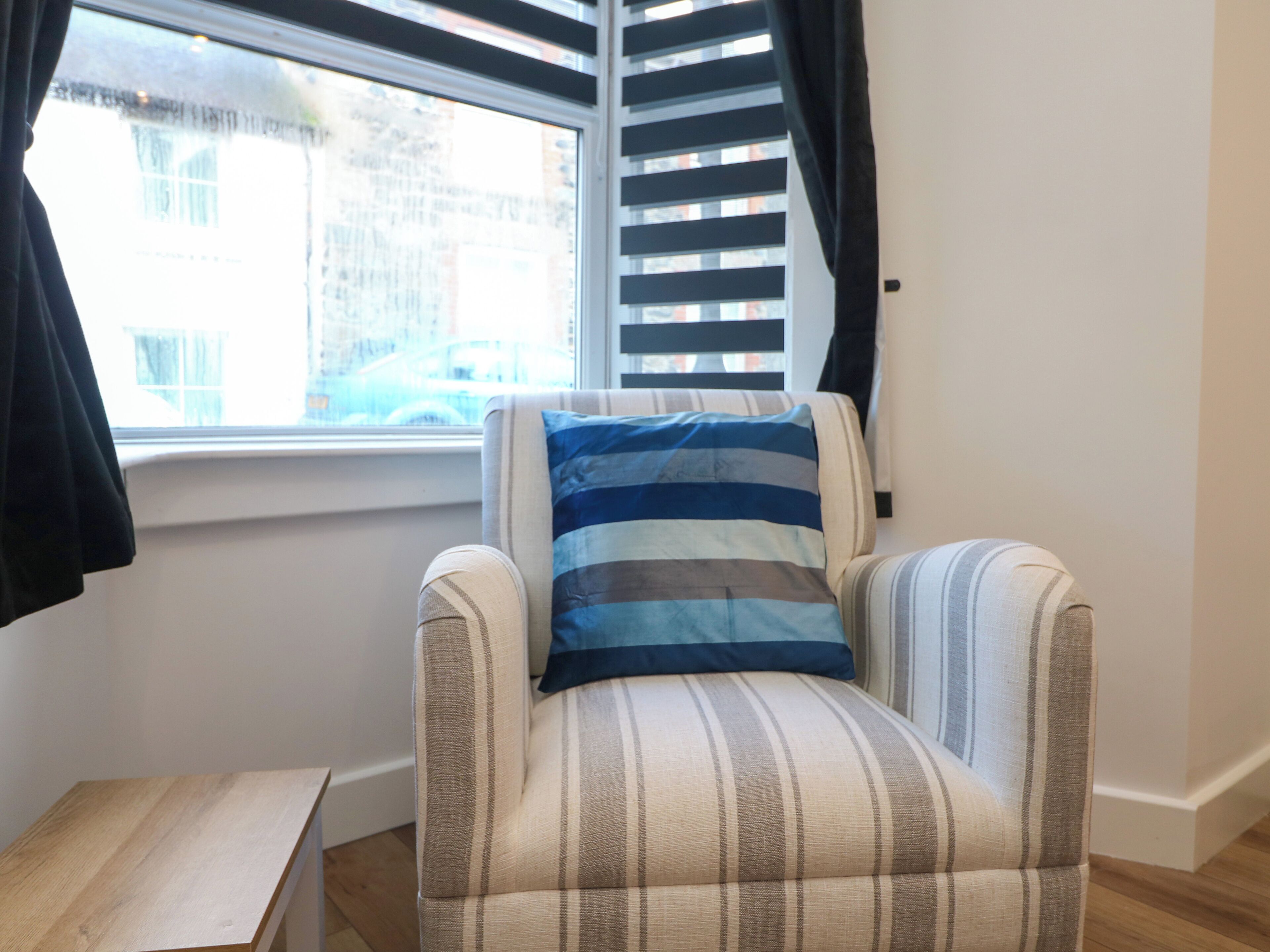 Ein Eckzimmer mit einem Streifenstuhl, einem kleinen Holztisch und einem Fenster mit Jalousien.
