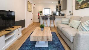 A modern living room with a TV, sofa, coffee table, and kitchen in the background. - Savoy Bay (Helston)