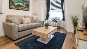 A modern living room with a beige sofa, a wooden coffee table, and a large painting of a coastal scene. - Savoy Bay (Helston)