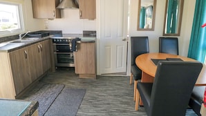 A compact kitchen with wooden cabinets, a stove, and a sink. There's a dining area with a table and chairs, and a door leading to another room. - 13 Gosling (Scarborough)