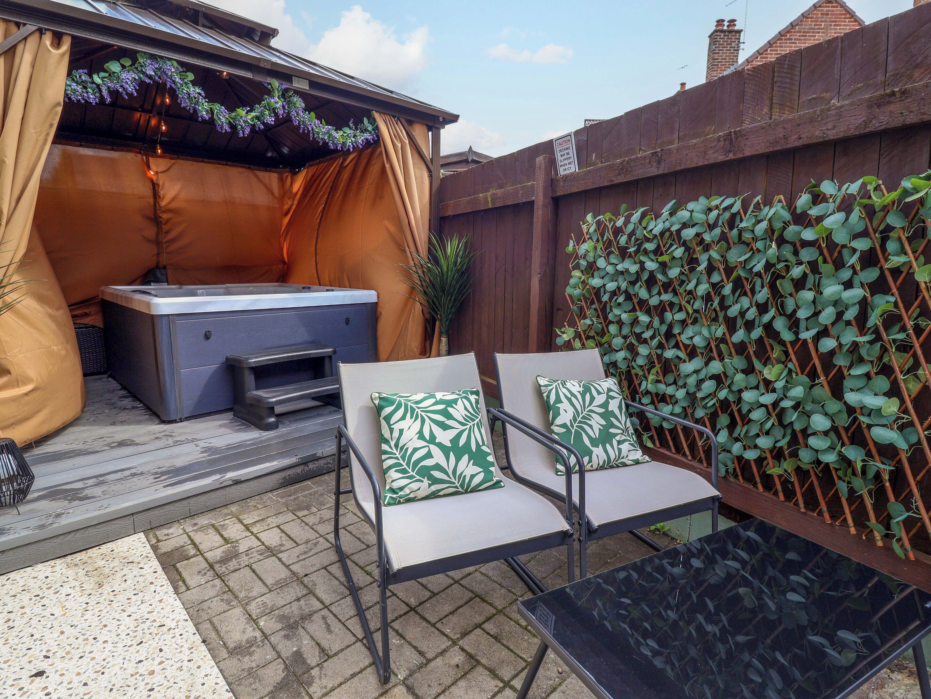 A patio with a hot tub, two chairs with cushions, a table, and a wooden fence with climbing plants.