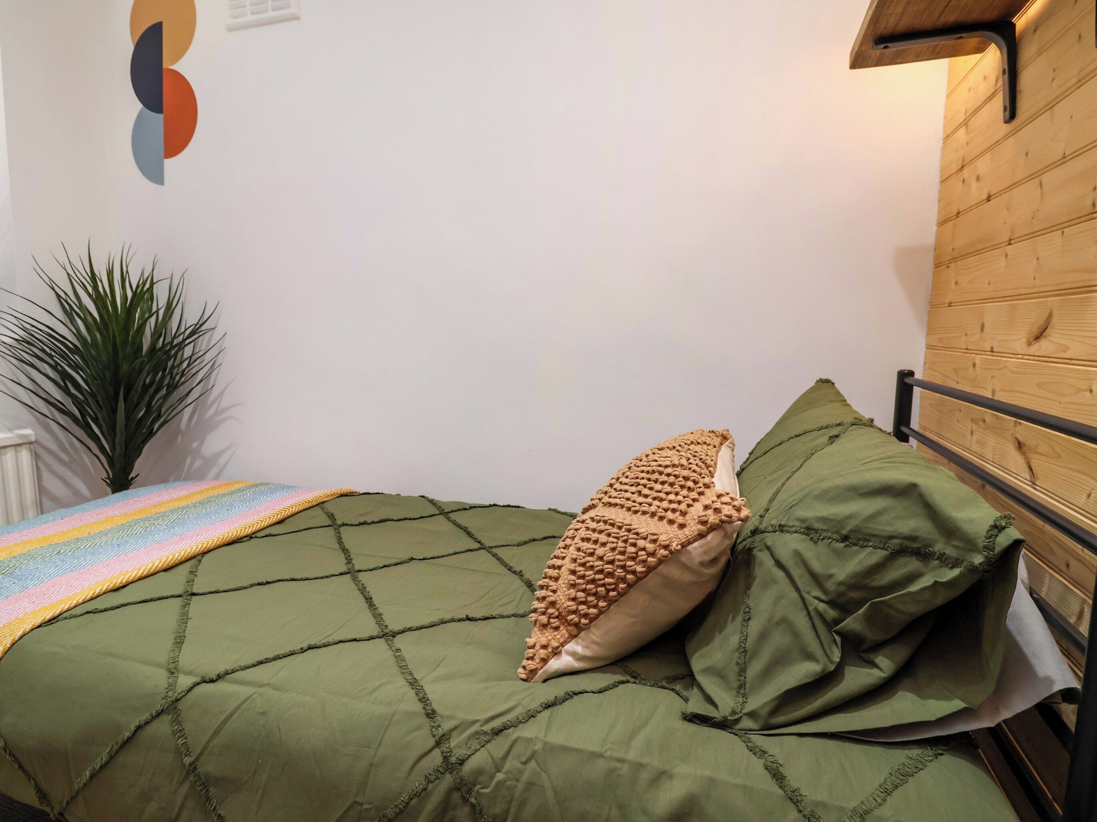 A bed with a green quilt, a single beige pillow, and a rainbow-striped blanket.