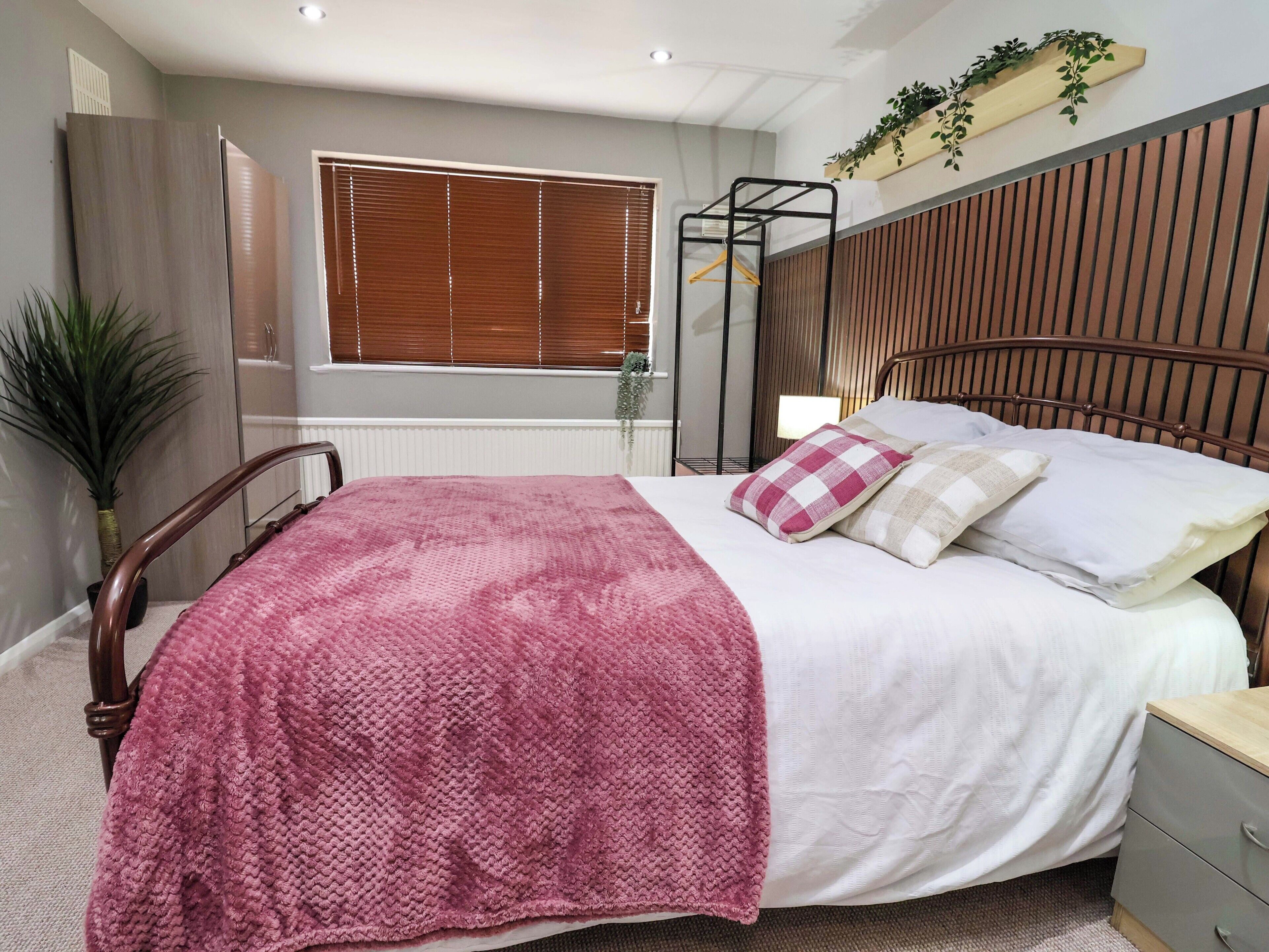 Une chambre à coucher comprenant un lit, une table de chevet, une commode, une fenêtre avec des stores, un radiateur, une plante en pot et une étagère murale agrémentée de plantes.