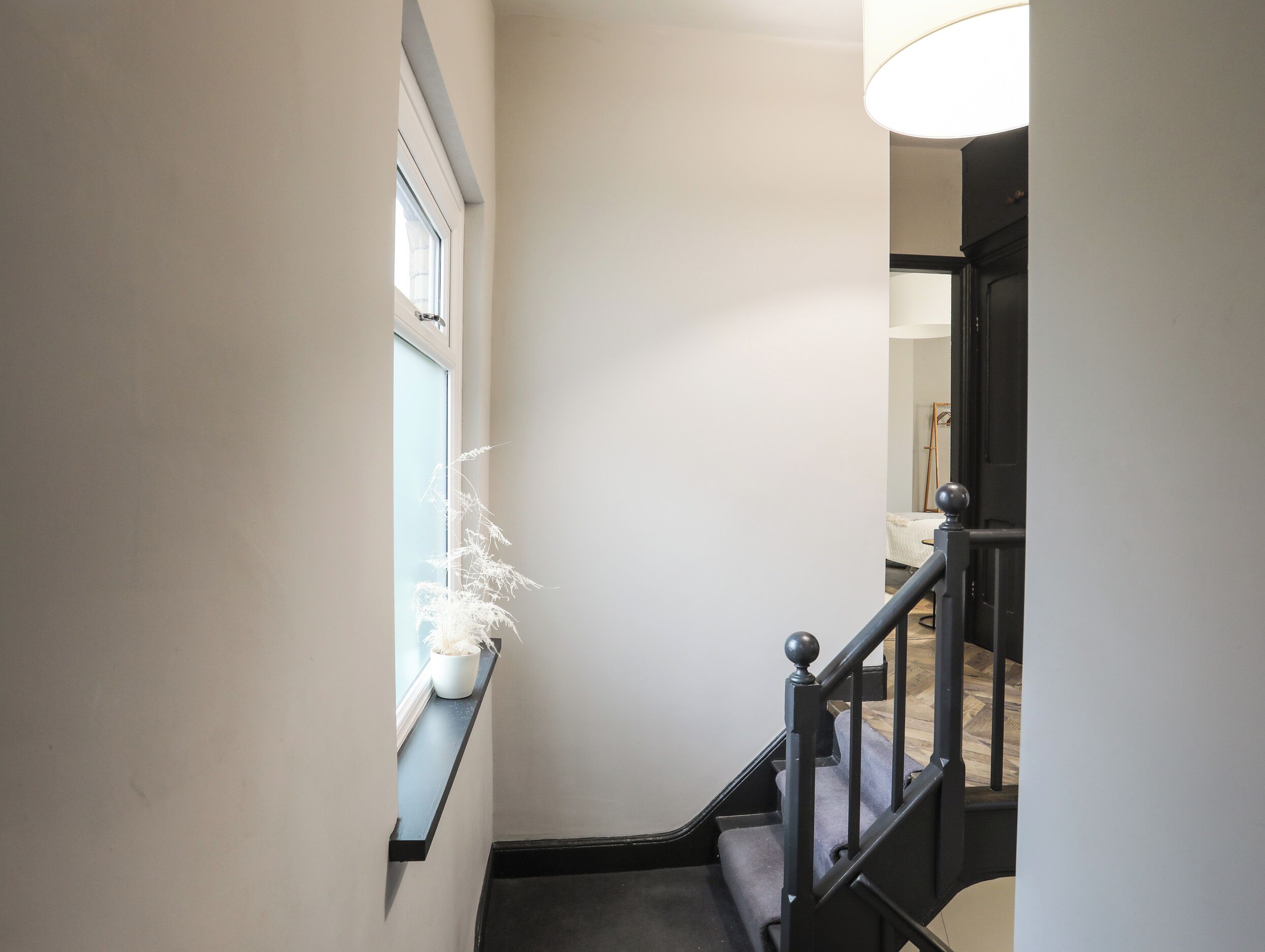 Un pasillo estrecho con una ventana a la izquierda, una escalera a la derecha y una puerta al final de la escalera.