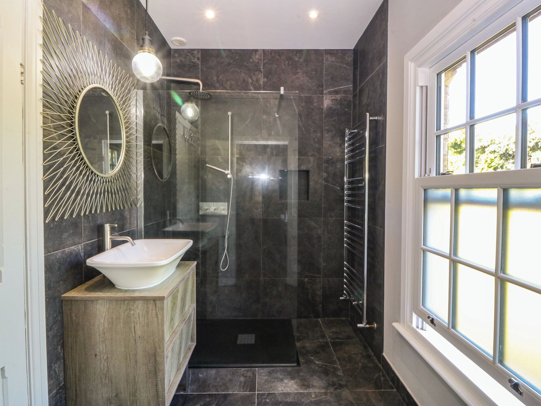 Un baño moderno con un lavamanos independiente, cabina de ducha con mampara de vidrio y un espejo grande con diseño de sol.