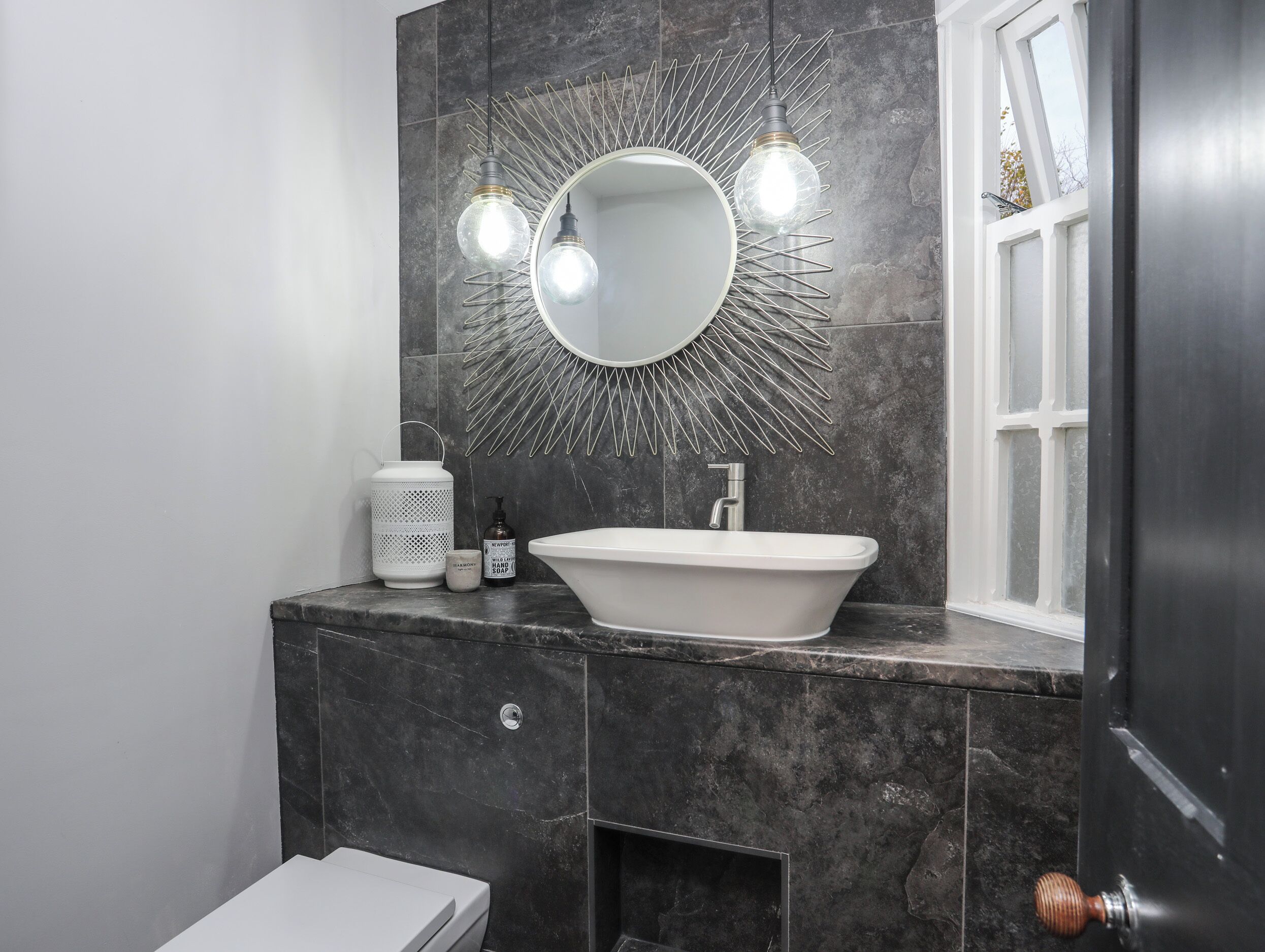 A modern bathroom with a freestanding bathtub, a round mirror with a sunburst light fixture, and a window with a view of the outside.