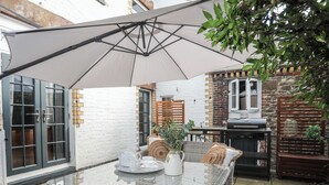 Une table à plateau en verre, entourée de chaises en osier, avec un pichet blanc et une théière, le tout placé sous un grand parasol dans une cour bordée d’un mur de briques et d’une clôture en bois.