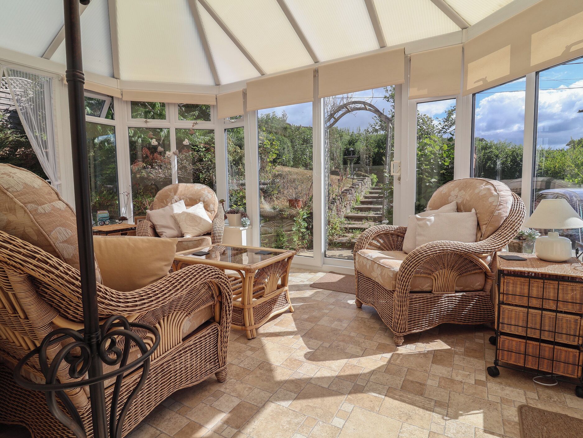 A sunlit room with wicker furniture, a glass table, and a view of a garden through large windows.